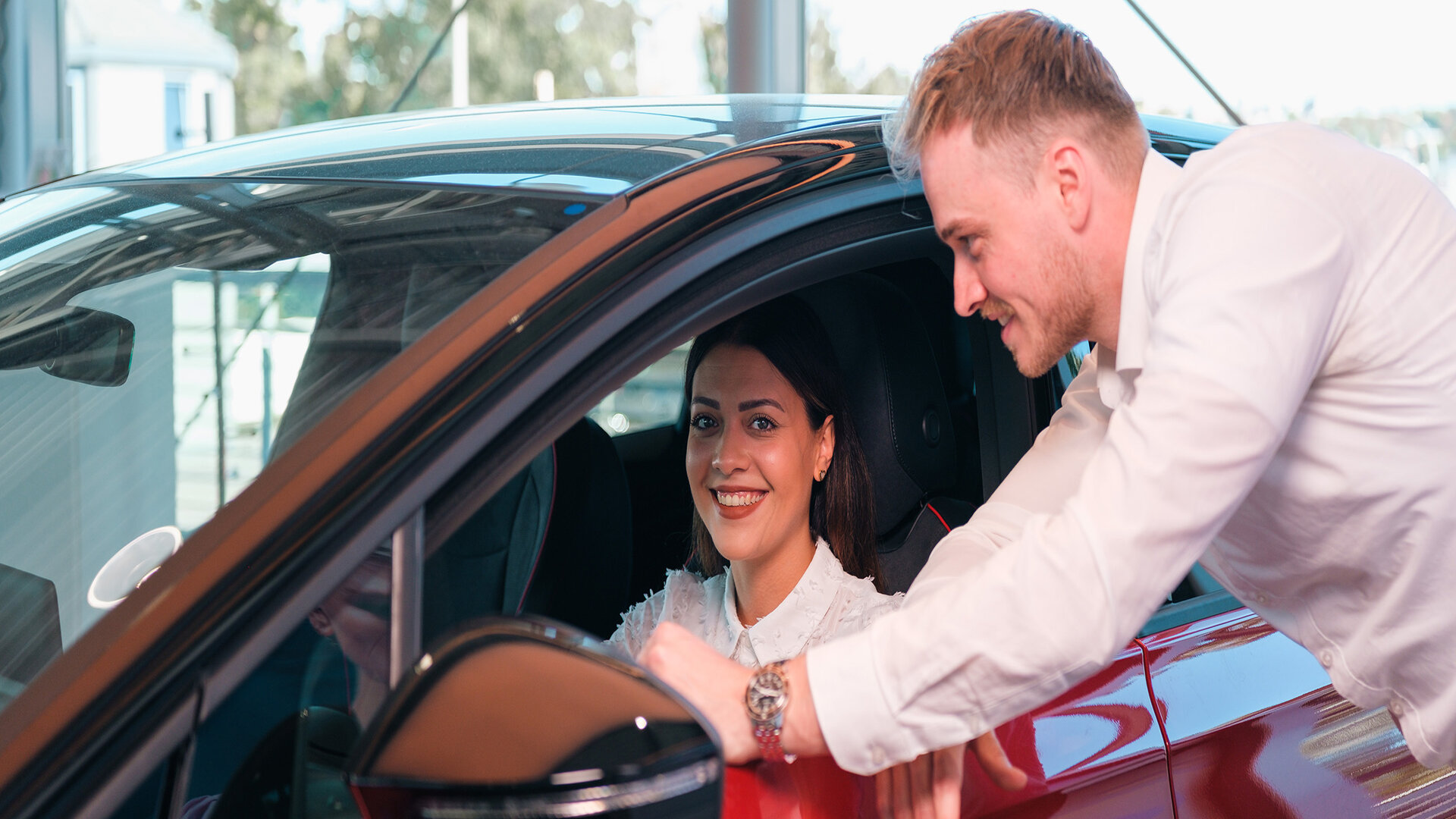 Auto Bach Alt-Text Generator sagte: Frau sitzt in rotem Fahrzeug im Auto Bach Autohaus, während ein Mitarbeiter am Fenster technische Details erklärt