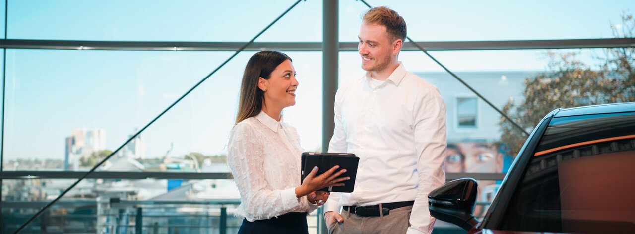 Zwei Mitarbeitende stehen im Autohaus neben einem Fahrzeug, sprechen miteinander und nutzen dabei ein Tablet.