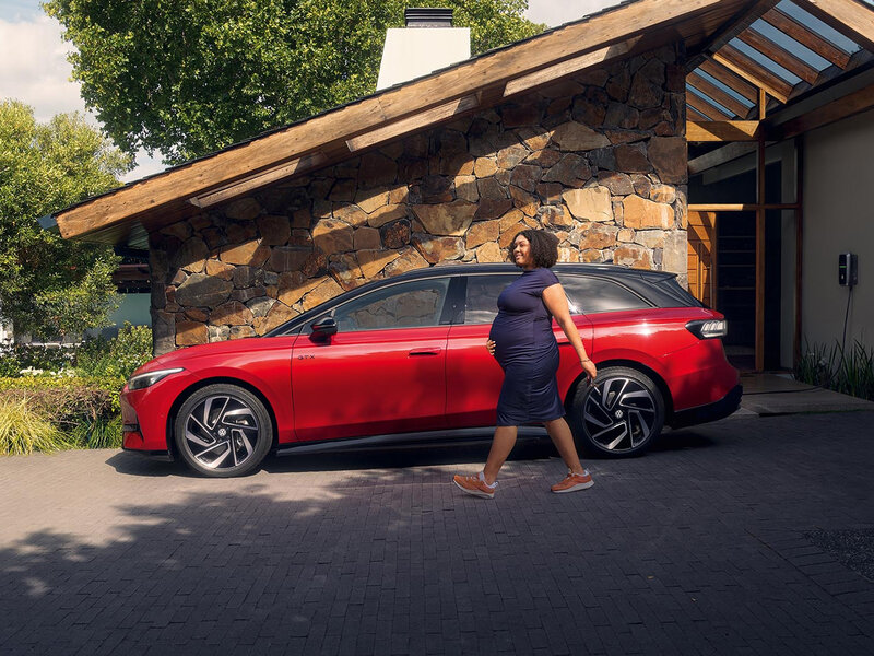 Frau geht am geparkten roten VW ID.7 GTX Tourer vorbei, modernes E-Kombi-Design vor architektonischem Haus.