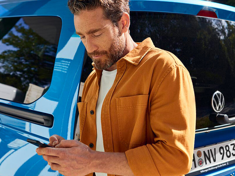 Eine Person steht neben einem geparkten Volkswagen Caddy und bedient ein Smartphone. Das Fahrzeug ist seitlich zu sehen, Fokus auf digitale Konnektivität im Alltag.