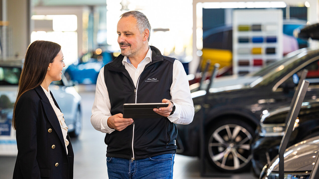 Beratungssituation im Autohaus: Ein Mitarbeiter zeigt einer Kundin Fahrzeugdetails auf einem Tablet, umgeben von ausgestellten Pkw.
