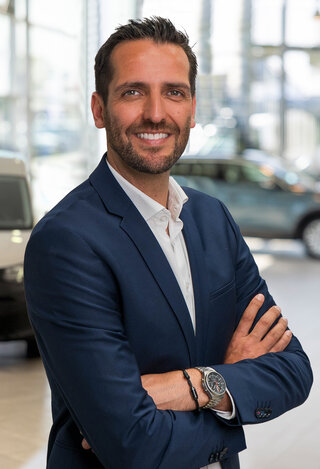 Sebastian Bach im blauen Anzug und weißen Hemd steht im Auto Bach Autohaus vor unscharfem Fahrzeughintergrund und blickt in die Kamera