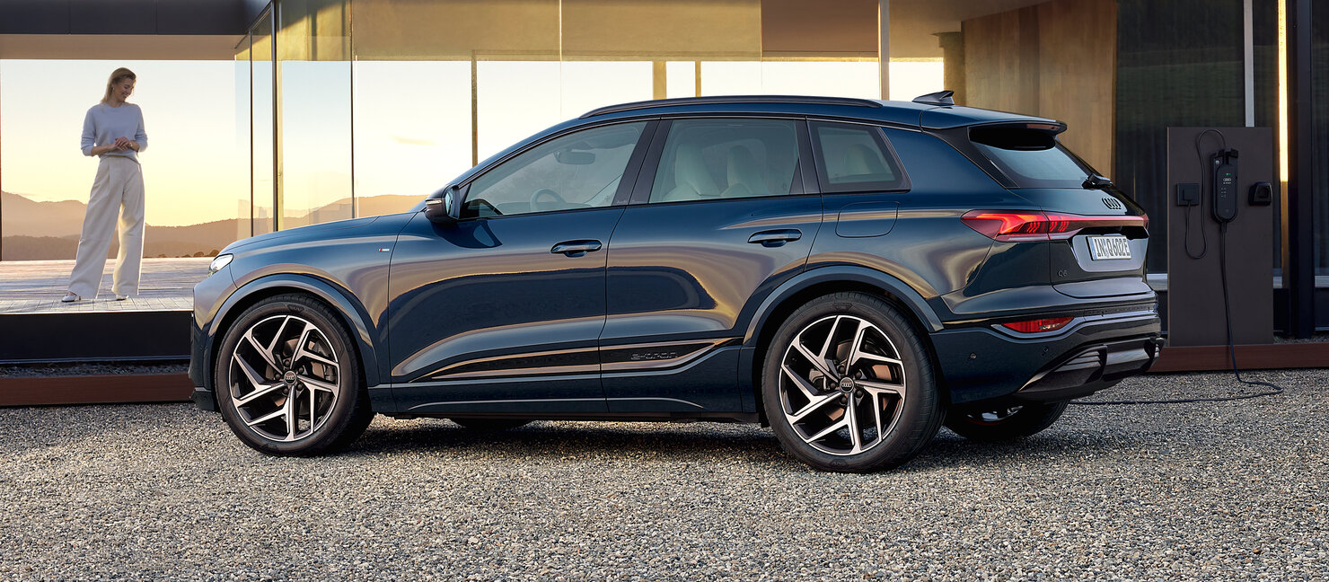 Audi Q6 e-tron seitlich geparkt an moderner Architektur mit Ladesäule, sportlich-elegantes SUV-Design und große Räder
