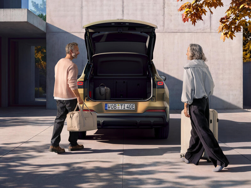 Goldfarbener Volkswagen ID. Tiguan mit geöffneter Heckklappe und zwei Personen mit Reisegepäck vor einem modernen Gebäude