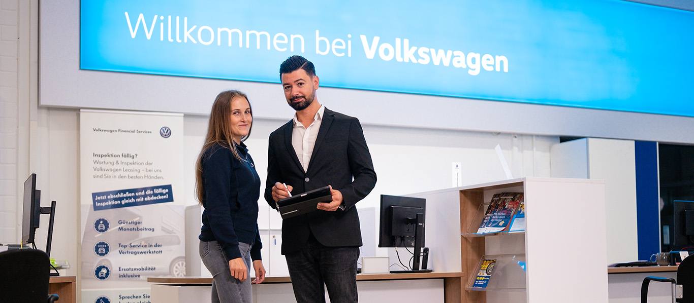 Zwei Personen stehen im Volkswagen Autohaus Auto Bach vor einem Empfangsbereich mit Tablet und Willkommensschild im Hintergrund