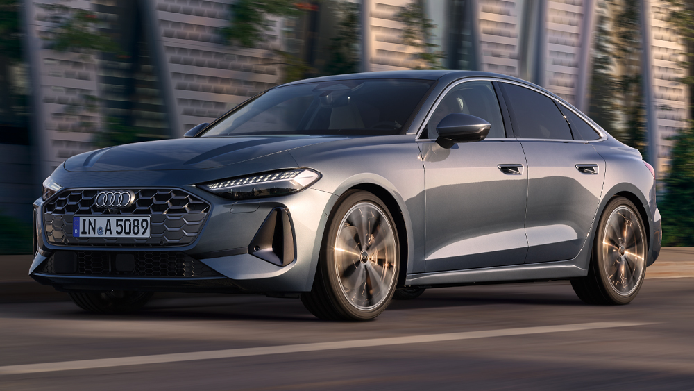 Audi A5 Limousine fährt dynamisch an moderner Architektur vorbei, schräger Frontblick bei Tageslicht