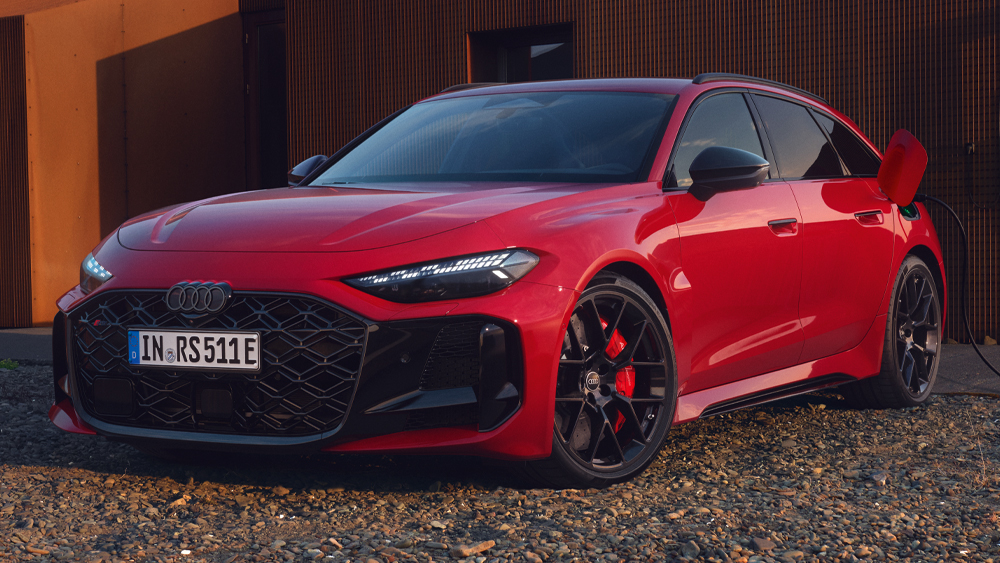 Roter Audi RS5 Avant in Frontansicht schräg von vorn auf Kiesfläche vor moderner Gebäudefassade bei Abendlicht