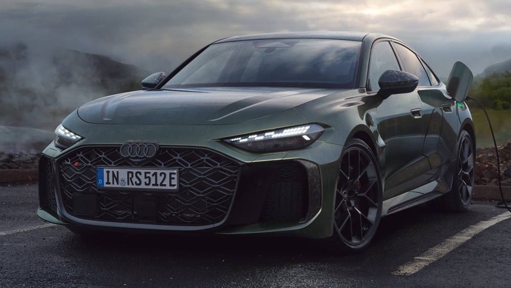 Audi RS5 Limousine in Frontansicht steht an einer Ladesäule bei bewölktem Himmel auf nasser Straße mit bergiger Landschaft im Hintergrund