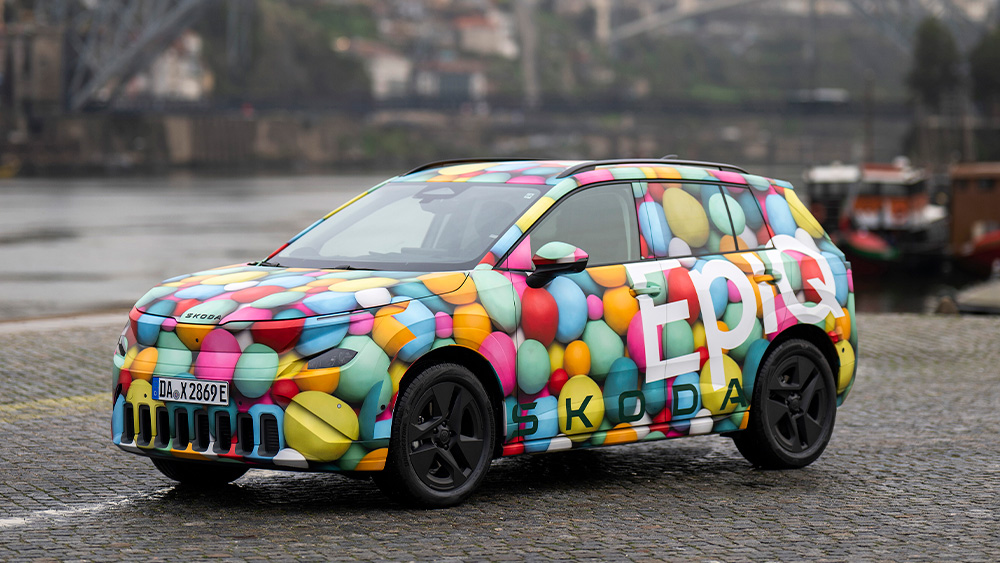 Bunt folierter Škoda Epiq steht auf gepflasterter Uferpromenade vor Fluss und Stahlbogenbrücke im Hintergrund, Seitenansicht schräg von vorn.