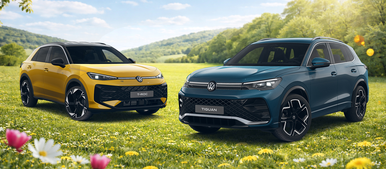 Volkswagen Tiguan und T-Roc stehen nebeneinander auf blühender Wiese vor hügeliger Landschaft unter blauem Himmel