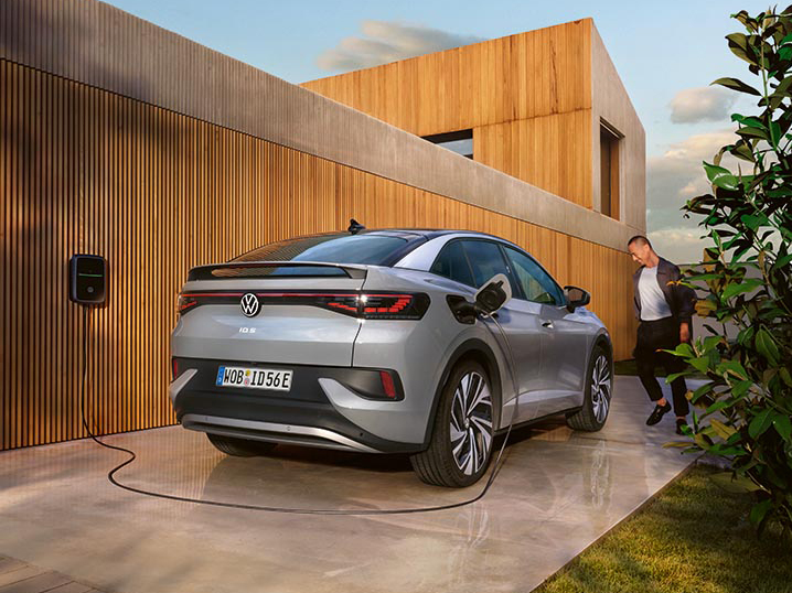 Grauer Volkswagen ID.5 steht vor einem modernen Haus und wird über eine Wallbox elektrisch geladen, eine Person geht am Fahrzeug vorbei.
