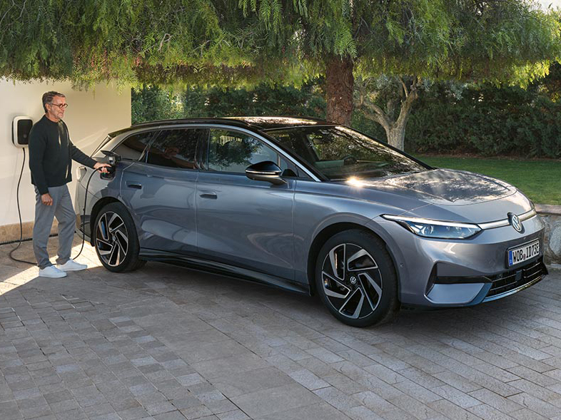Grauer VW ID.7 Tourer steht vor einem Haus und wird an einer Wallbox geladen, umgeben von Garten und Tageslicht.