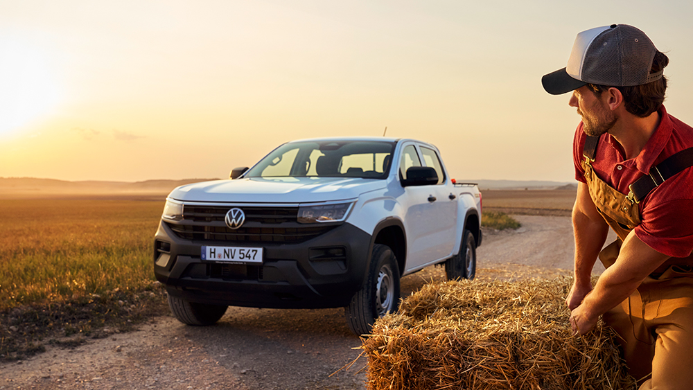 Weißer Volkswagen Amarok steht bei Sonnenuntergang auf Feldweg, Person sitzt auf Heuballen daneben.