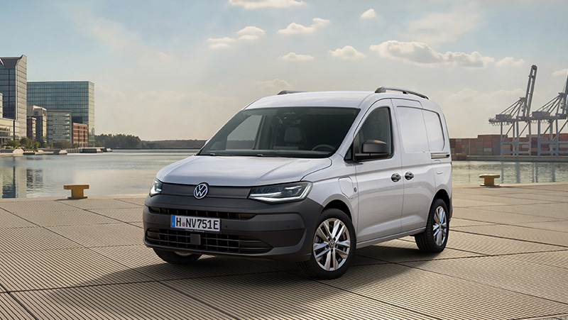 Volkswagen Caddy Kastenwagen in Frontansicht parkt auf einer Hafenpromenade mit Wasser, Containerkränen und moderner Stadtarchitektur im Hintergrund