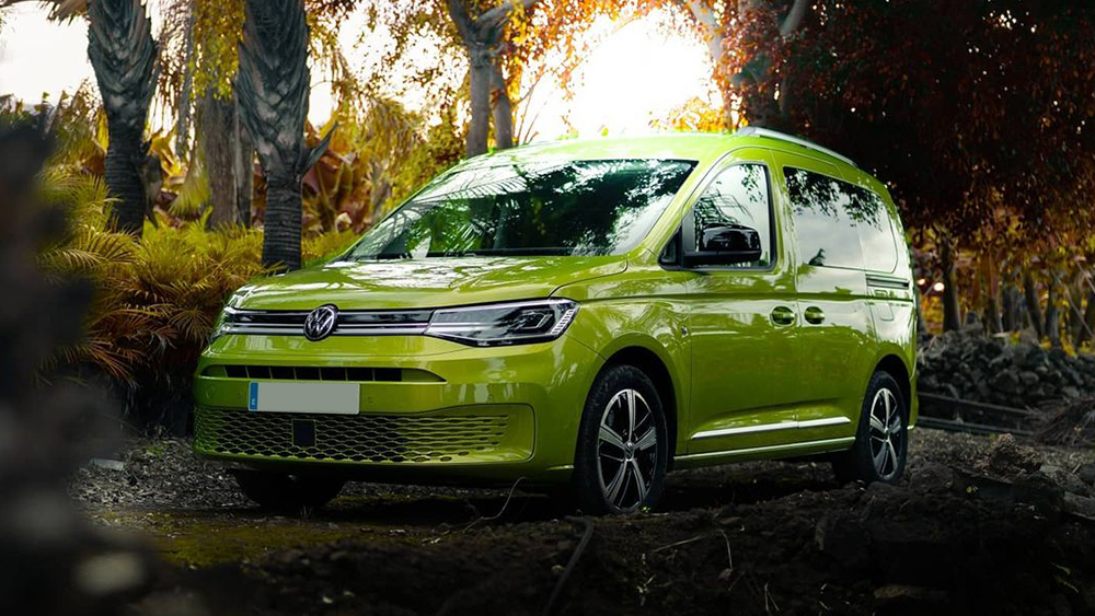 Grüner Volkswagen Caddy fährt durch herbstlichen Wald, Sonnenlicht fällt durch Bäume.