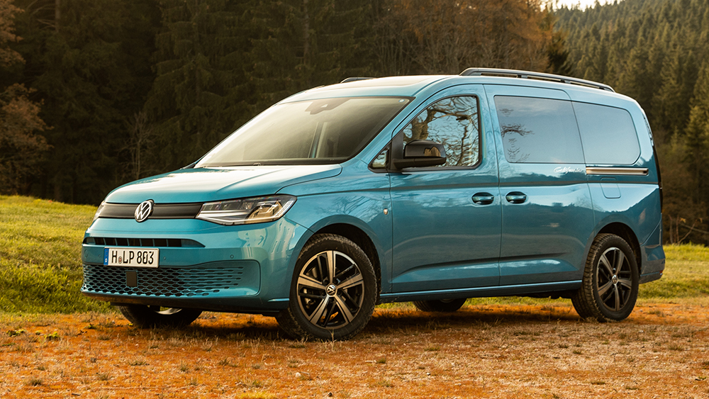 Blauer Volkswagen Caddy California steht auf Waldlichtung im Herbst, umgeben von Bäumen und Laub.
