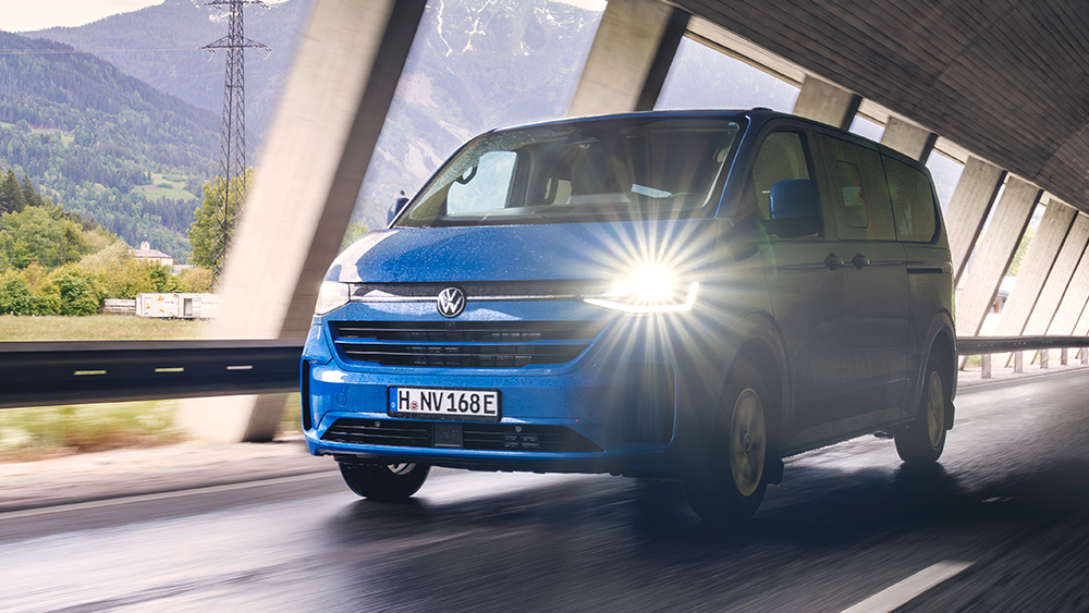 Blauer Volkswagen Caravelle fährt durch Tunnel, Scheinwerfer leuchtet, Berge im Hintergrund.