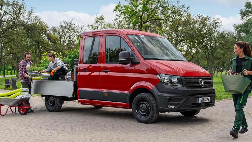 Drei Personen transportieren Pflanzen und Arbeitsmaterial zu einem roten Crafter-Pritschenwagen auf einem Parkweg.