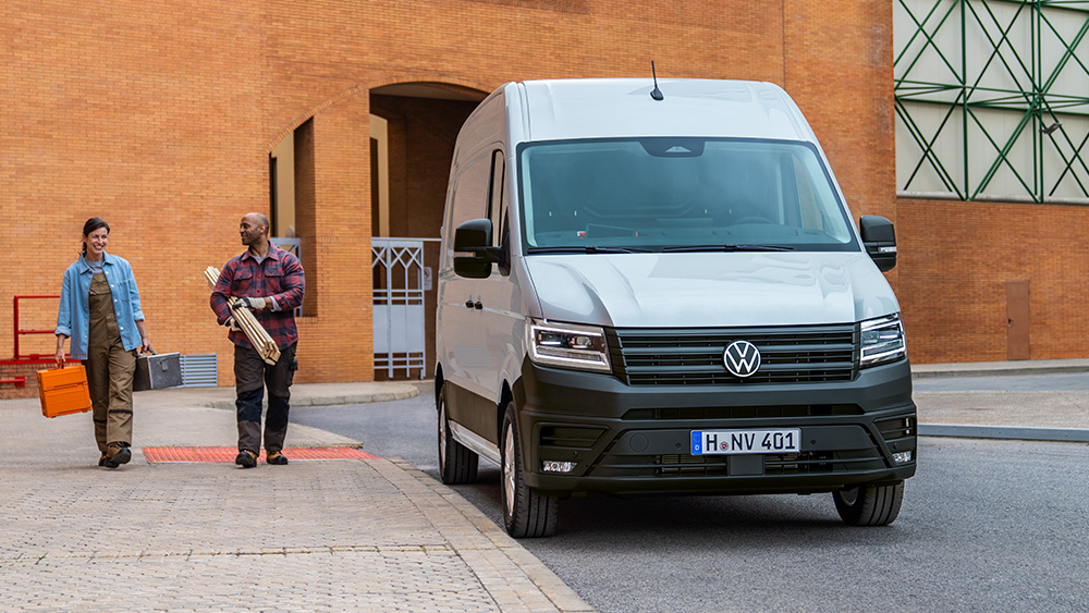 Zwei Personen gehen mit Koffern auf einen weißen Transporter zu, der vor einem Gebäude parkt.