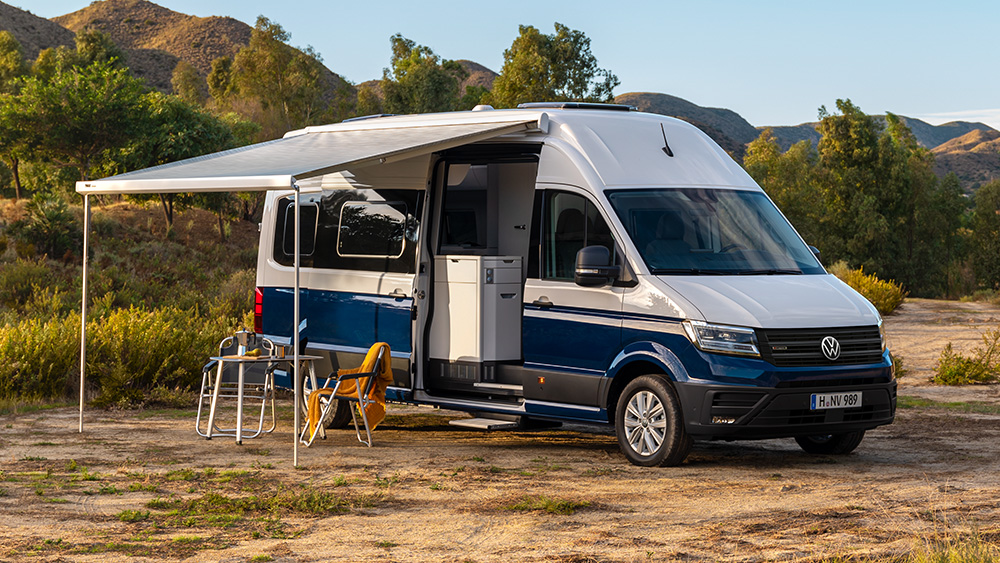 Volkswagen Grand California Camper steht in Natur, Markise ausgefahren, Stühle und Tisch davor.