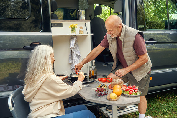 Volkswagen Grand California Camper mit ausgefahrener Küche, an dem zwei Personen im Freien Essen zubereiten und an einem Klapptisch sitzen