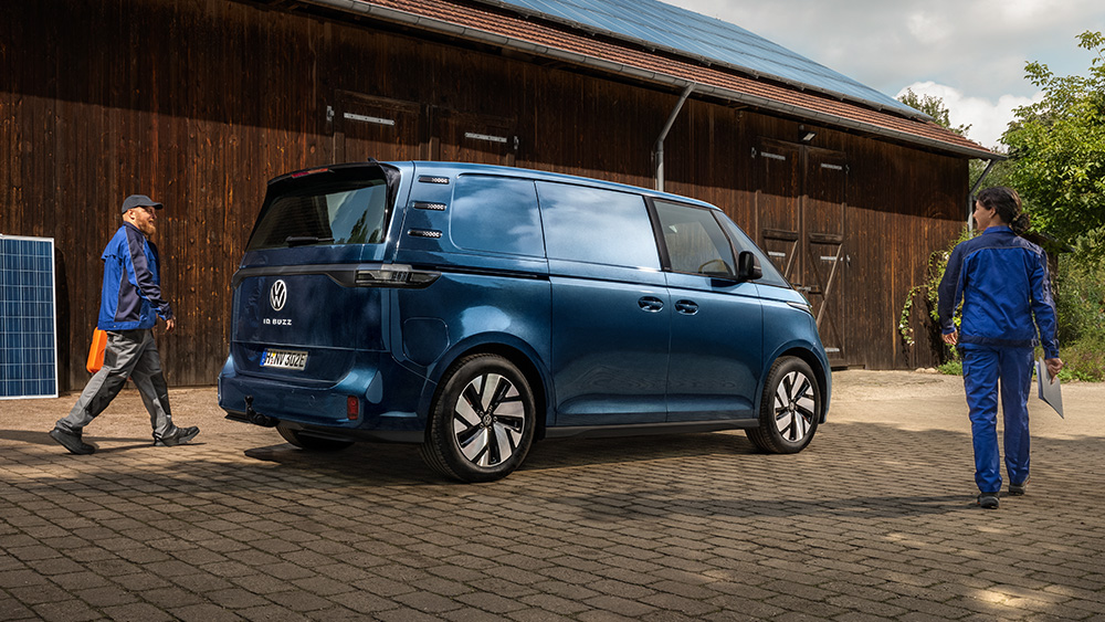 Blauer Volkswagen ID. Buzz Cargo steht vor Werkhalle, zwei Personen in Arbeitskleidung gehen daneben.