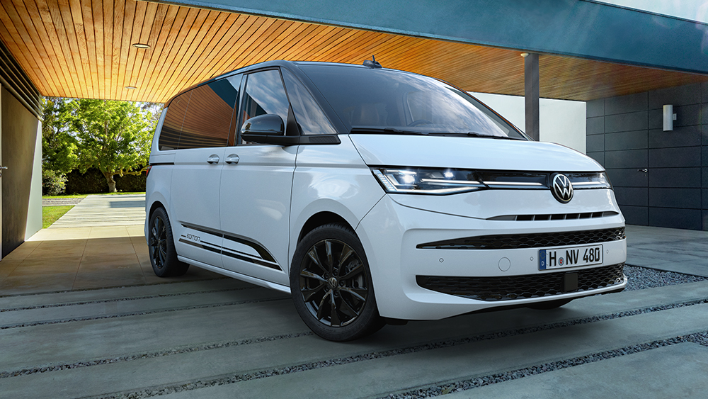 Weißer Volkswagen Multivan steht vor modernem Holzgebäude, klare Architektur und Tageslicht.