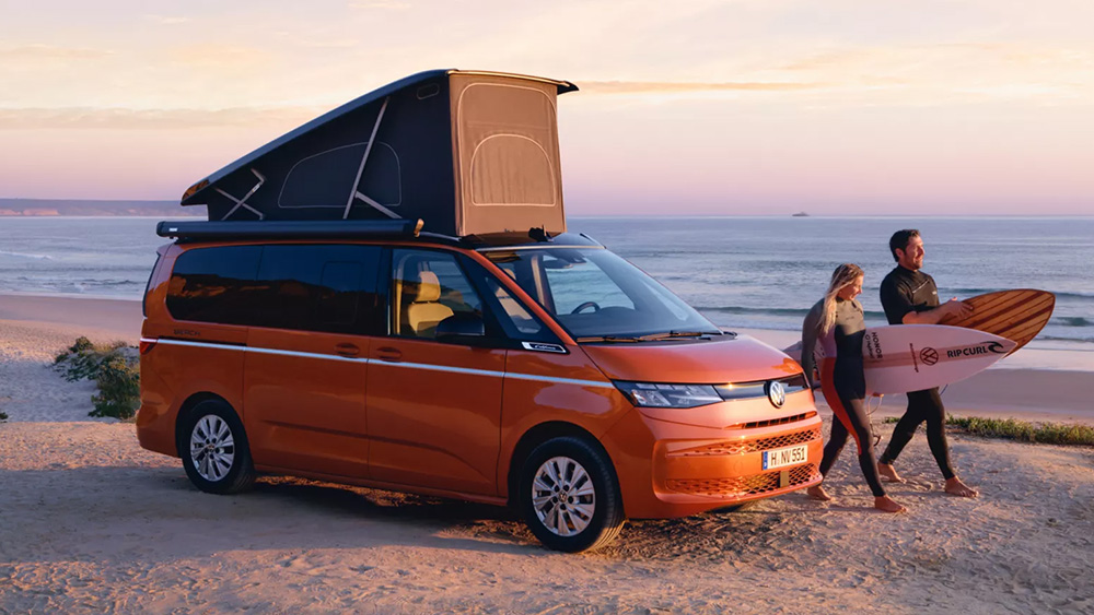 Oranger Volkswagen California mit geöffnetem Aufstelldach steht am Strand bei Sonnenuntergang. Daneben gehen zwei Personen barfuß über den Sand, eine davon trägt ein Surfbrett, im Hintergrund ist das Meer zu sehen.