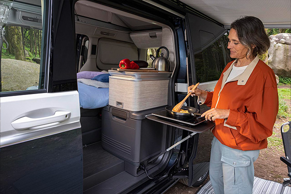 Person bereitet im Innenraum eines Volkswagen California eine Mahlzeit in der integrierten Küche des Fahrzeugs zu.